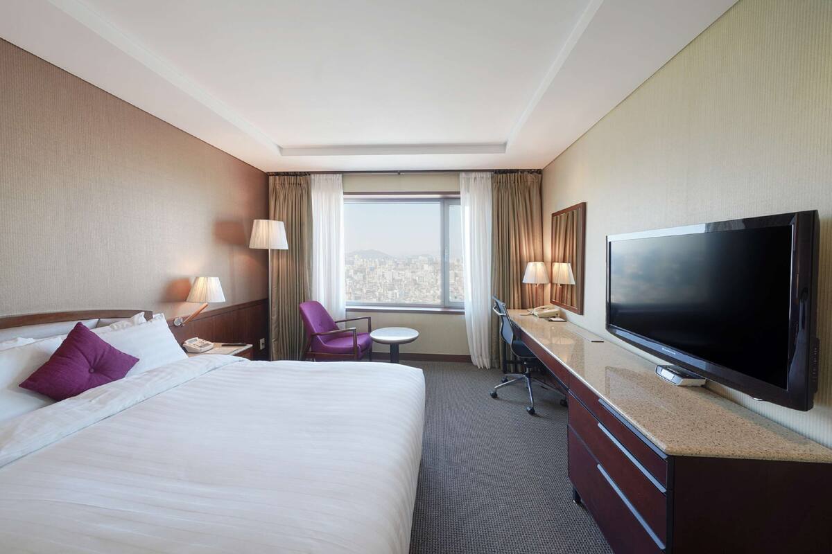 Een hotelkamer met een groot bed, een bureau met een flatscreen televisie, een stoel, een klein tafeltje en een raam met uitzicht op de stad.