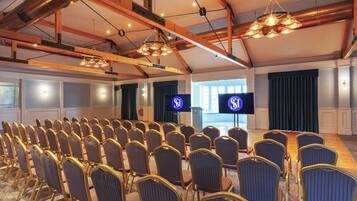 Un salón de conferencias con filas de sillas, un escenario con un atril y dos pantallas mostrando el logo.