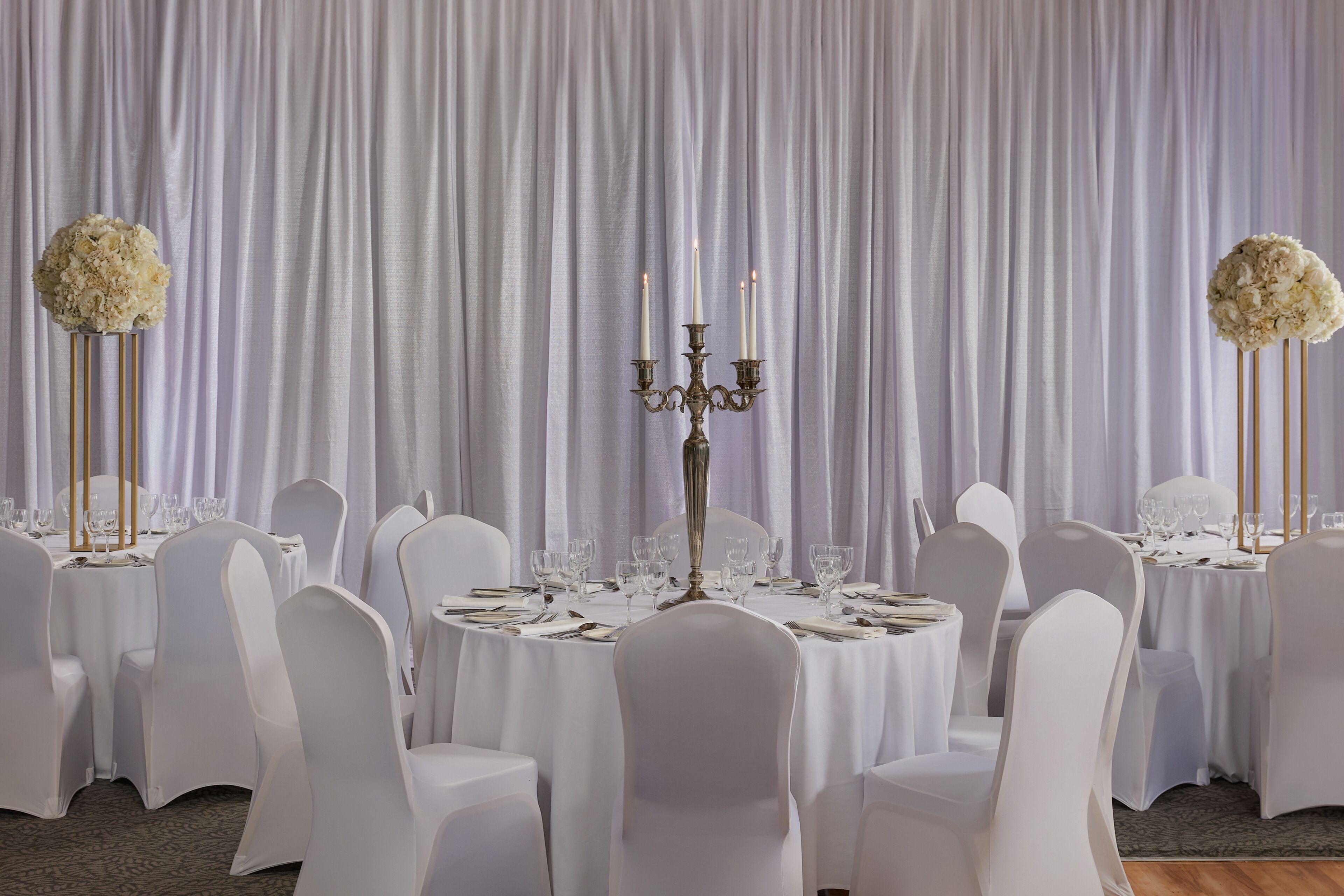 A banquet hall with round tables set for dining, white chairs, and a backdrop of white curtains.
