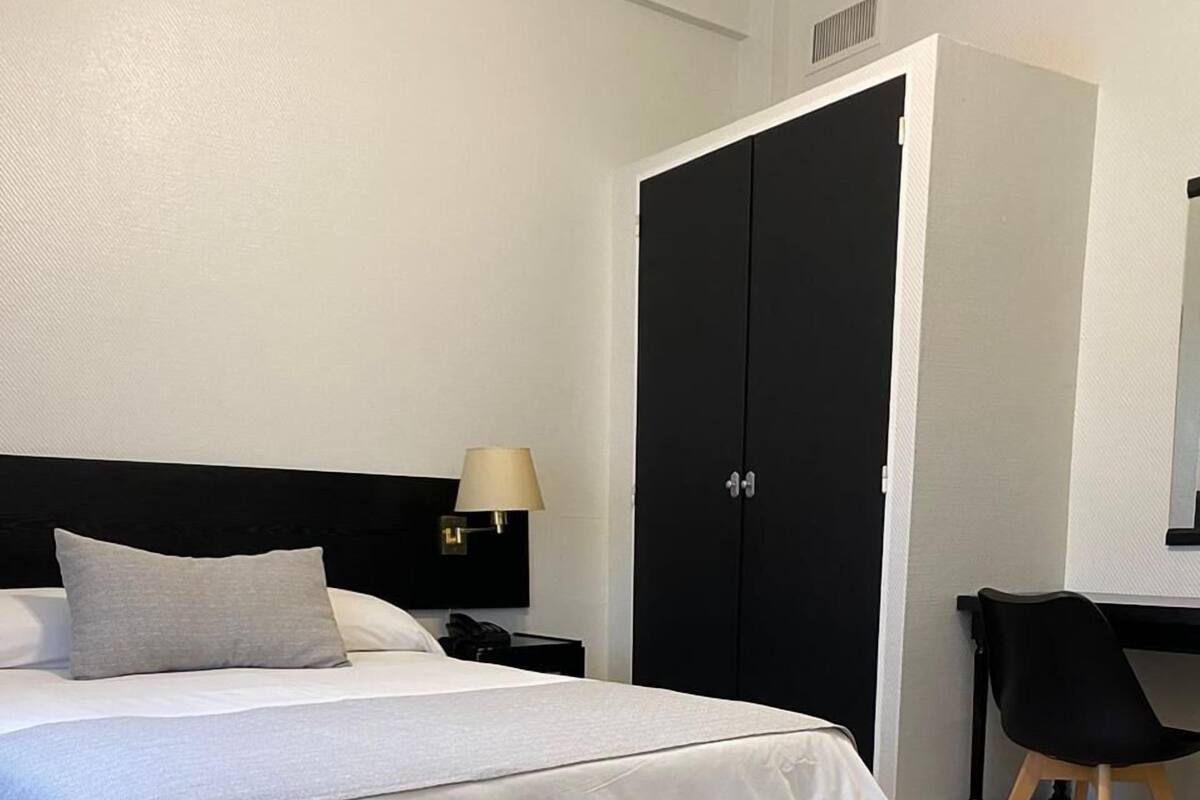 A neatly made bed with a grey pillow, a bedside table with a lamp, a black wardrobe, and a wooden chair.