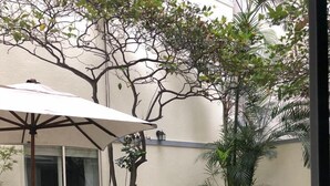 Un área de patio al aire libre con mesas y sillas redondas, una jardinera central con plantas verdes y un paraguas blanco que da sombra.