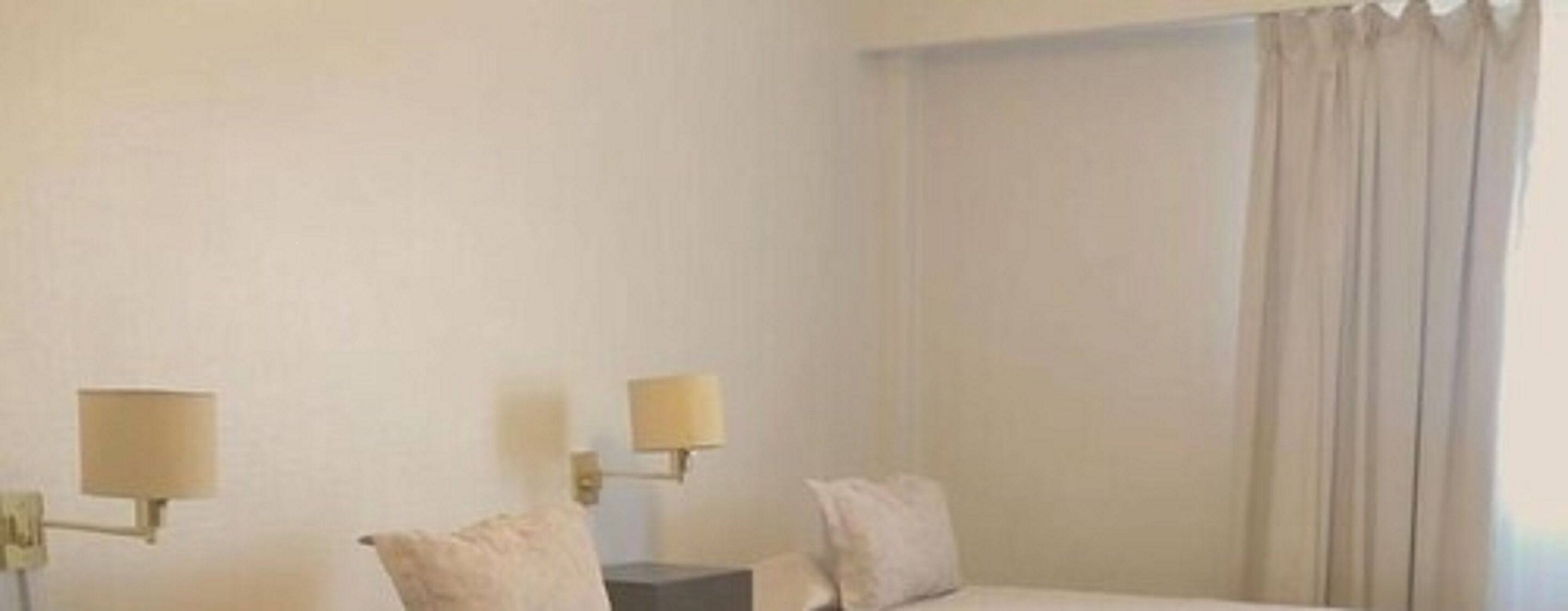 A hotel room with a bed, two bedside lamps, a nightstand, and a window with curtains.