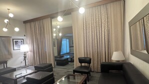 Una habitación de hotel moderna con una sala de estar que incluye un sofá, otomanas, una mesa de centro de vidrio y una araña.