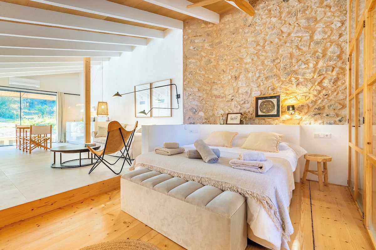 A bedroom with a stone wall, a bed with multiple pillows, a bench, and a small stool.