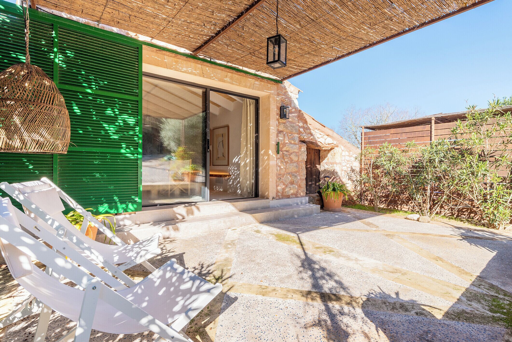 Un patio con suelo de piedra, una silla lounge blanca, una silla colgante de mimbre y una puerta con contraventanas verdes.
