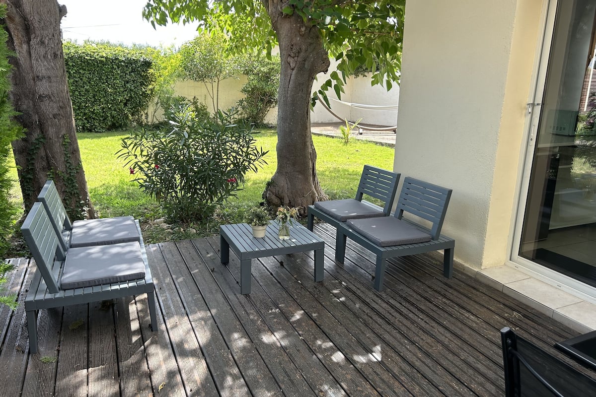 A wooden deck with two grey benches and a table, surrounded by greenery and trees.