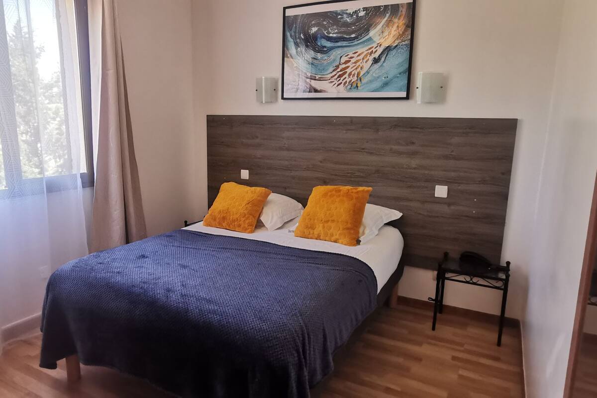 A bedroom with a bed, two orange pillows, a blue blanket, a wooden headboard, a painting above the bed, and a bedside table with a phone.