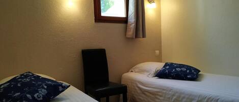 A small, single-room accommodation with two beds, each with a pillow and a chair.