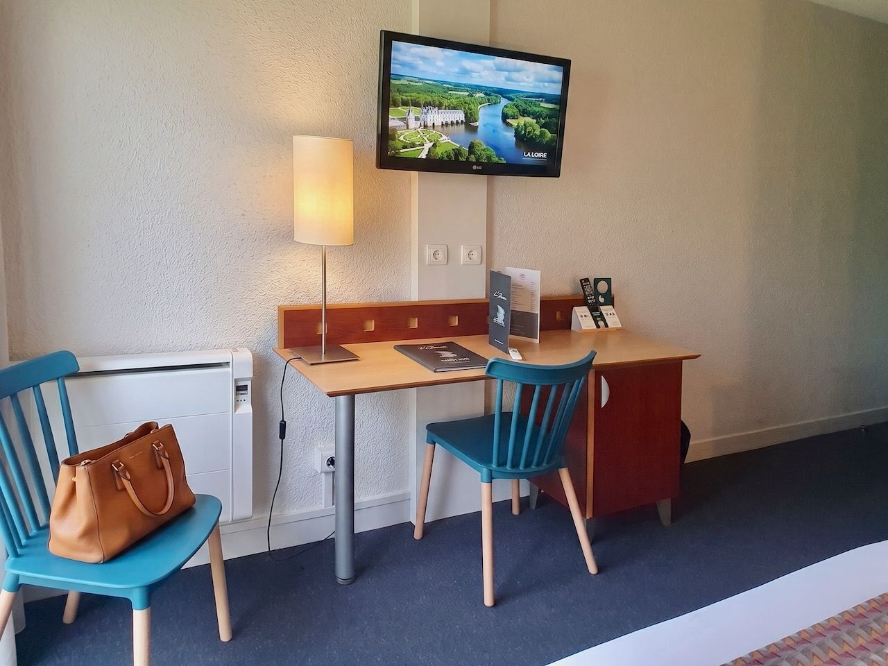 A hotel room with a desk, two chairs, a television mounted on the wall, a lamp, and a brown handbag on a chair.