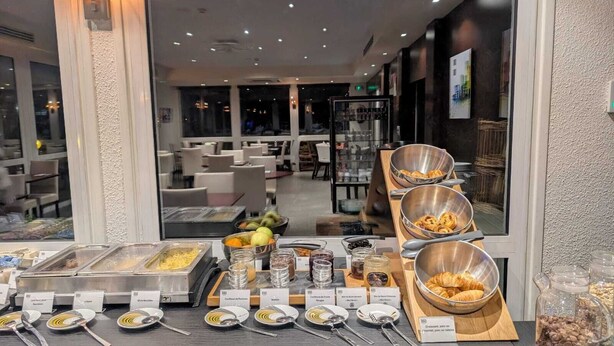 Un bufé con diversos alimentos y utensilios, un área de estar con mesas y sillas, y vistas al exterior a través de amplias ventanas.
