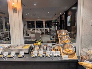 Un buffet proposant une variété de plats et d’ustensiles, un espace repas avec des tables et des chaises, et une vue sur l’extérieur grâce à de grandes fenêtres.