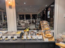 Buffet com diversos alimentos e utensílios, área de refeições com mesas e cadeiras, e vista para o exterior através de grandes janelas.