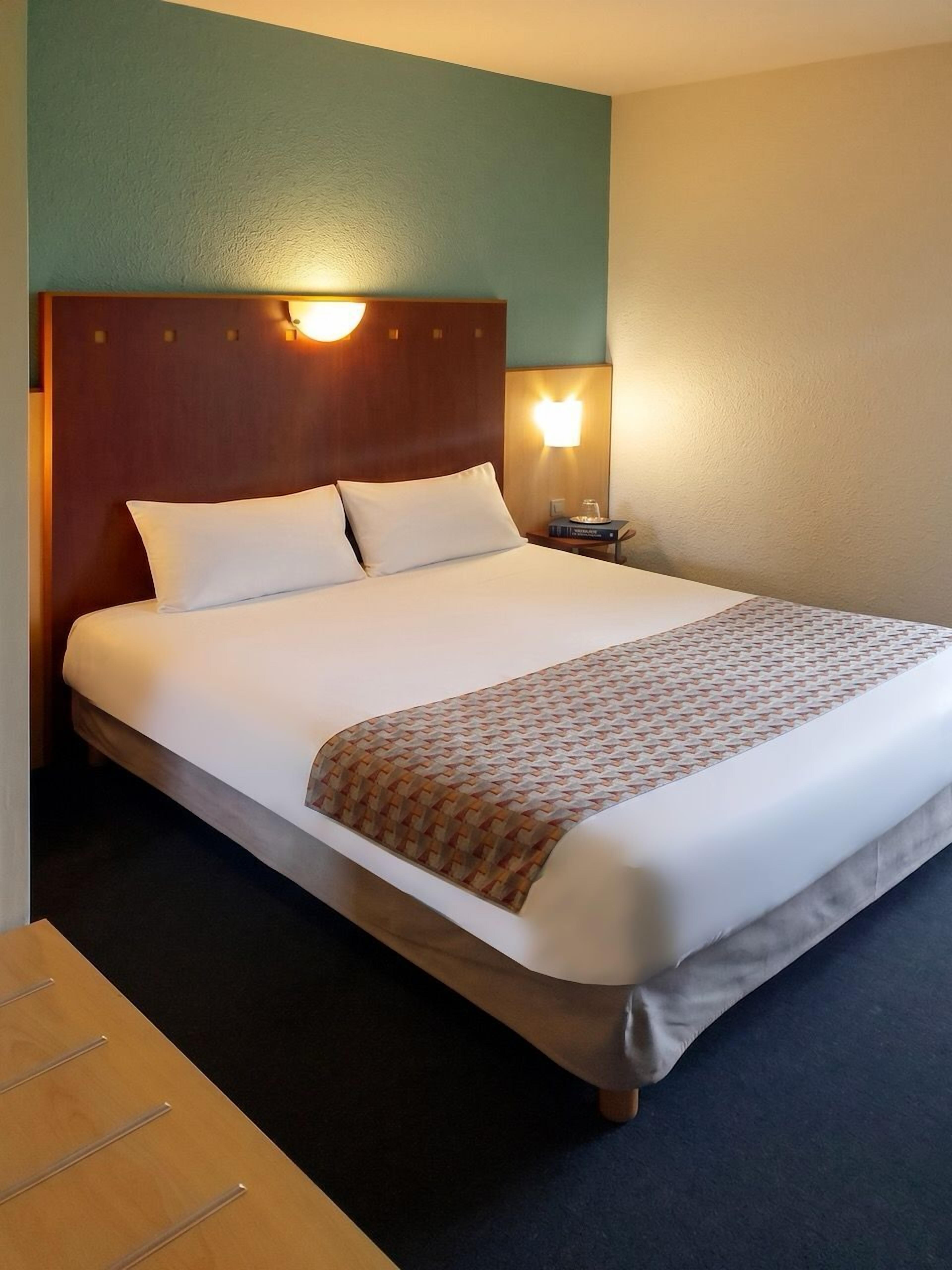 A hotel room with a bed, a nightstand, and a wall-mounted lamp.