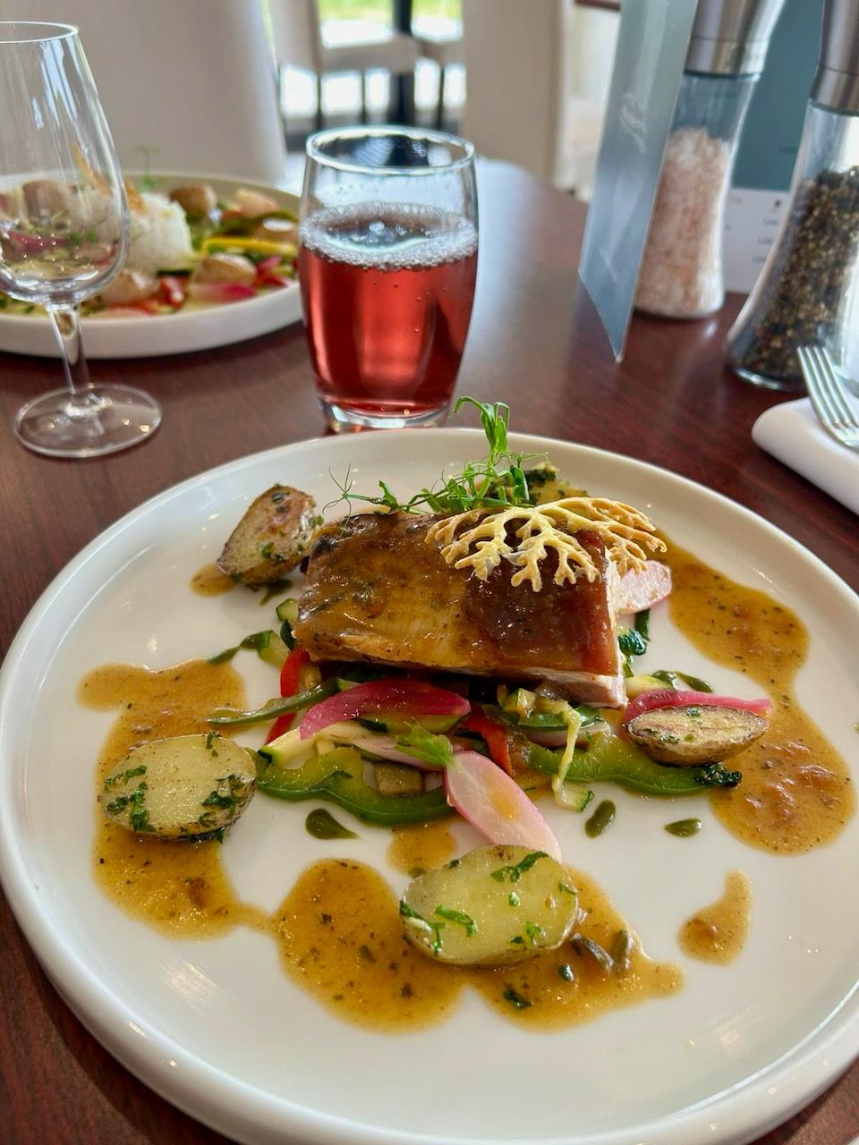 A plate of food with a fish fillet, vegetables, and sauce, accompanied by a glass of red beverage and a glass of white beverage.