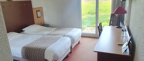 A hotel room with two single beds, a desk with a lamp, a chair, and a view of a grassy area through the window.