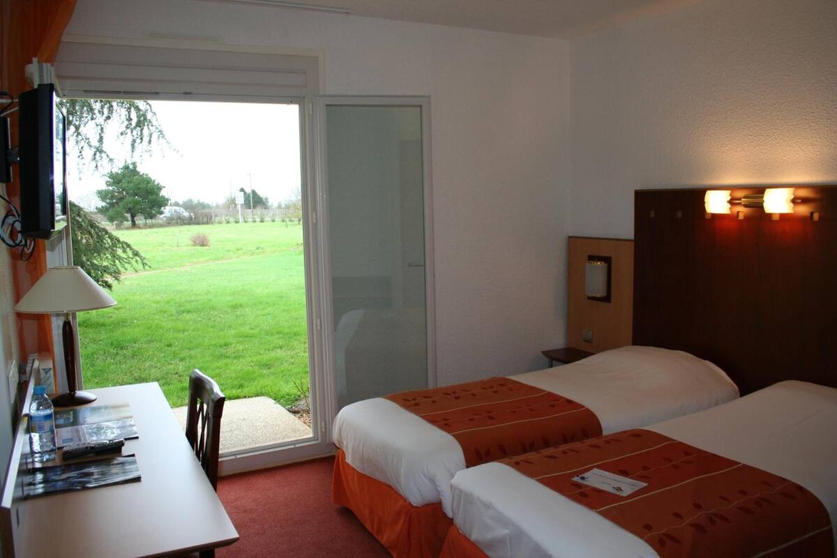 Hotelkamer met twee bedden, een bureau met stoel, een televisie en uitzicht op een gazon door een schuifdeur.
