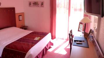 A hotel room with a bed, a desk with a lamp, a chair, a TV mounted on the wall, and a window with curtains.