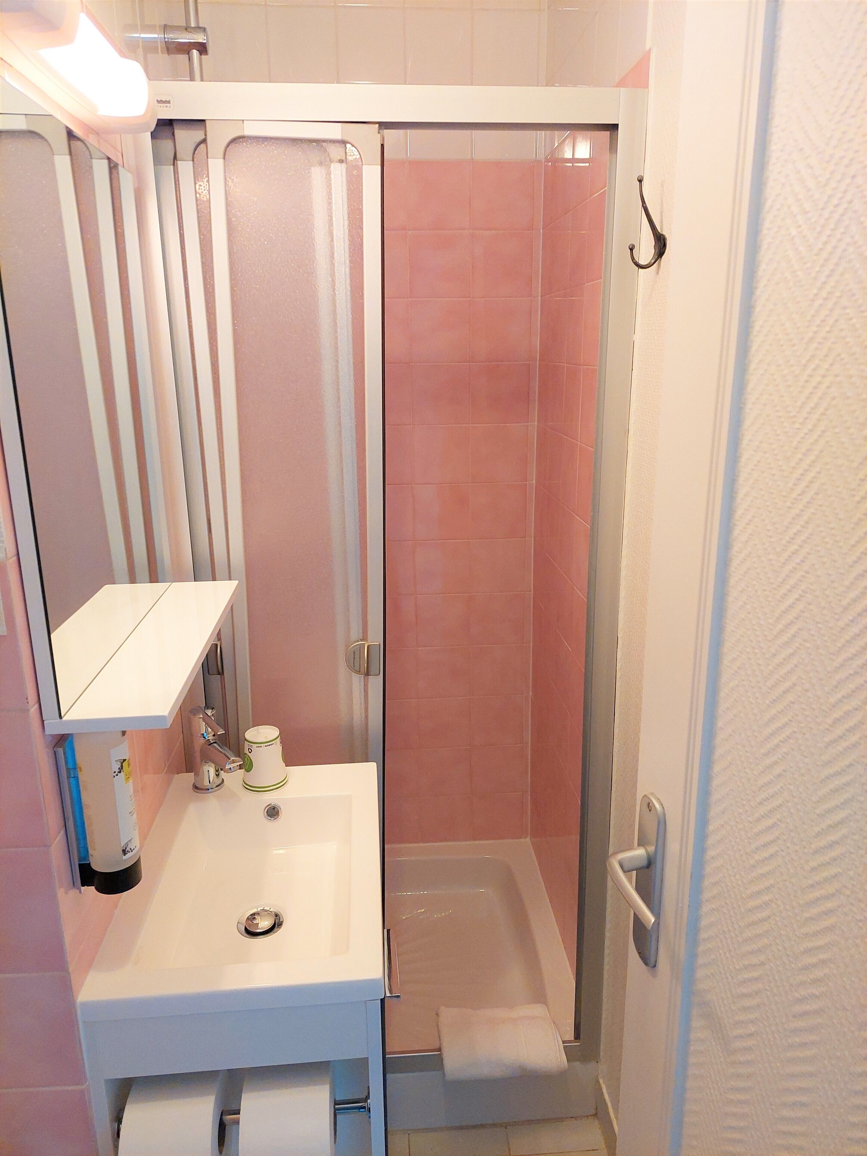 Ein Badezimmer mit weißem Waschbecken, einem Spiegel, einem Handtuchhalter und einer Dusche mit rosa Kacheln.