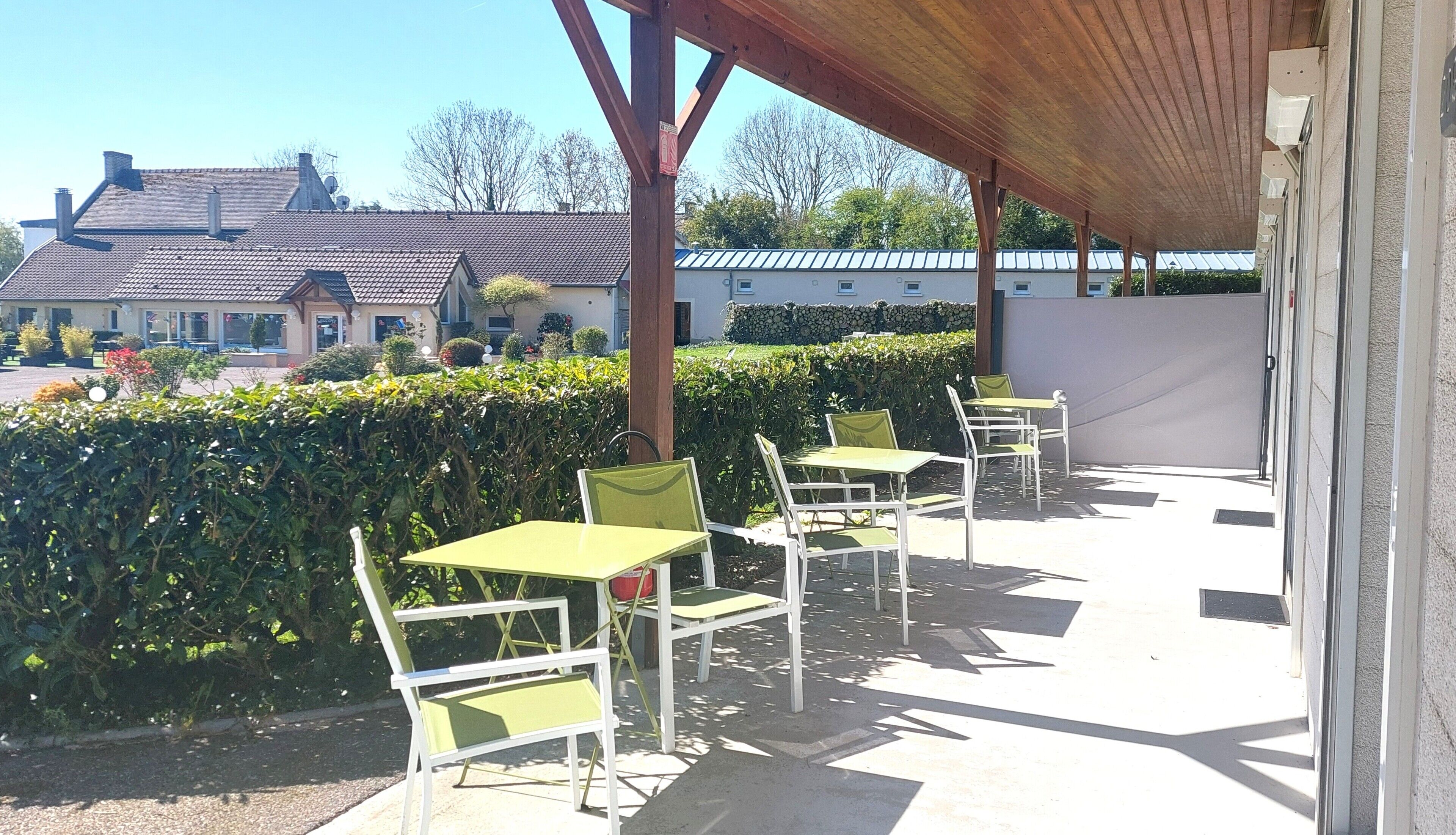Un espace extérieur abrité, avec des tables et des chaises, une haie et des bâtiments en arrière-plan.