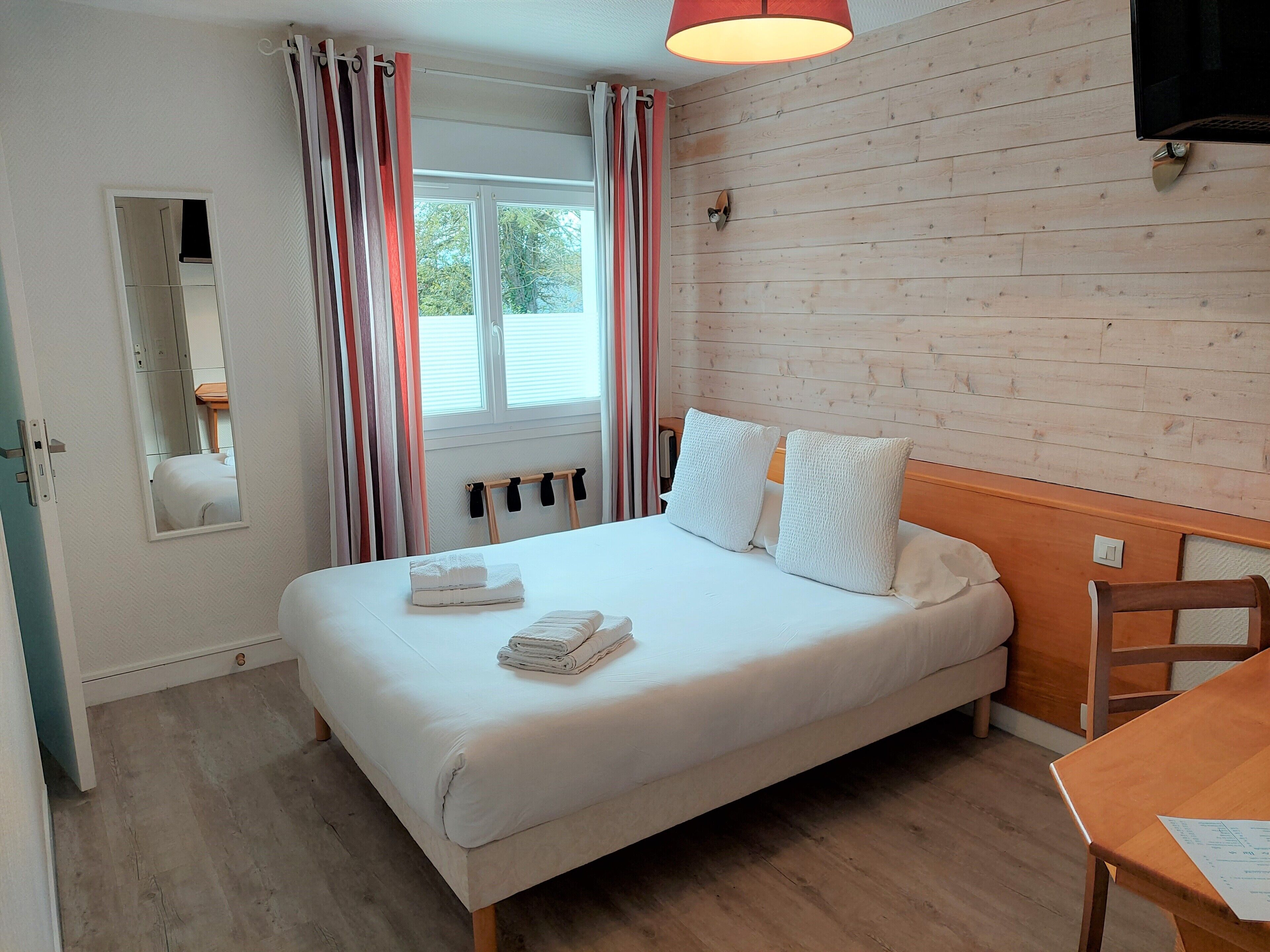 Una cama bien hecha con almohadas y toallas dobladas, un cabecero de madera, una mesa de madera con un menú y una ventana con cortinas.