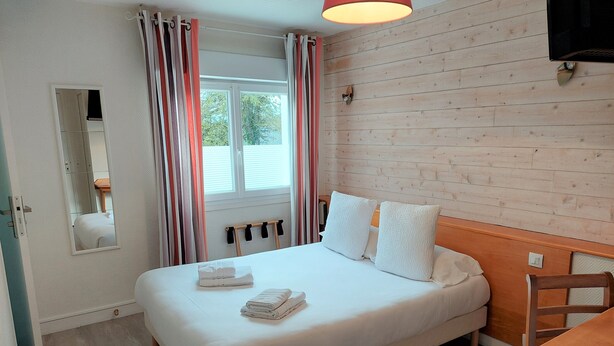 Una cama bien hecha con almohadas y toallas dobladas, un cabecero de madera, una mesa de madera con un menú y una ventana con cortinas.