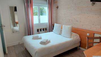 Una cama bien hecha con almohadas y toallas dobladas, un cabecero de madera, una mesa de madera con un menú y una ventana con cortinas.