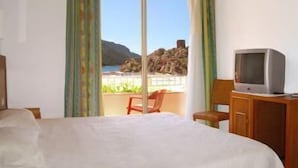 A hotel room with a bed, a television on a wooden cabinet, and a view of the beach and mountains through a large sliding door. - Hotel Kalliste (Ota)