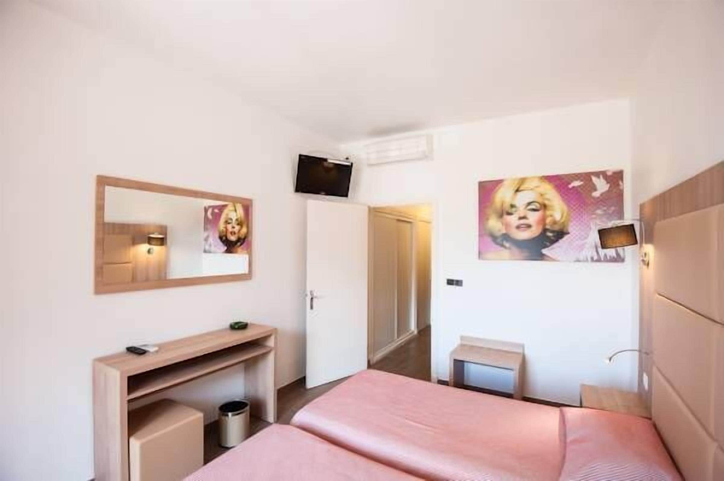 Habitación de hotel con cama, escritorio, espejo, televisión y obras de arte en las paredes.