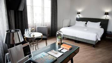 Quarto de hotel com cama, luminárias de cabeceira, escrivaninha com luminária, uma cadeira, uma mesinha e janela com cortinas.