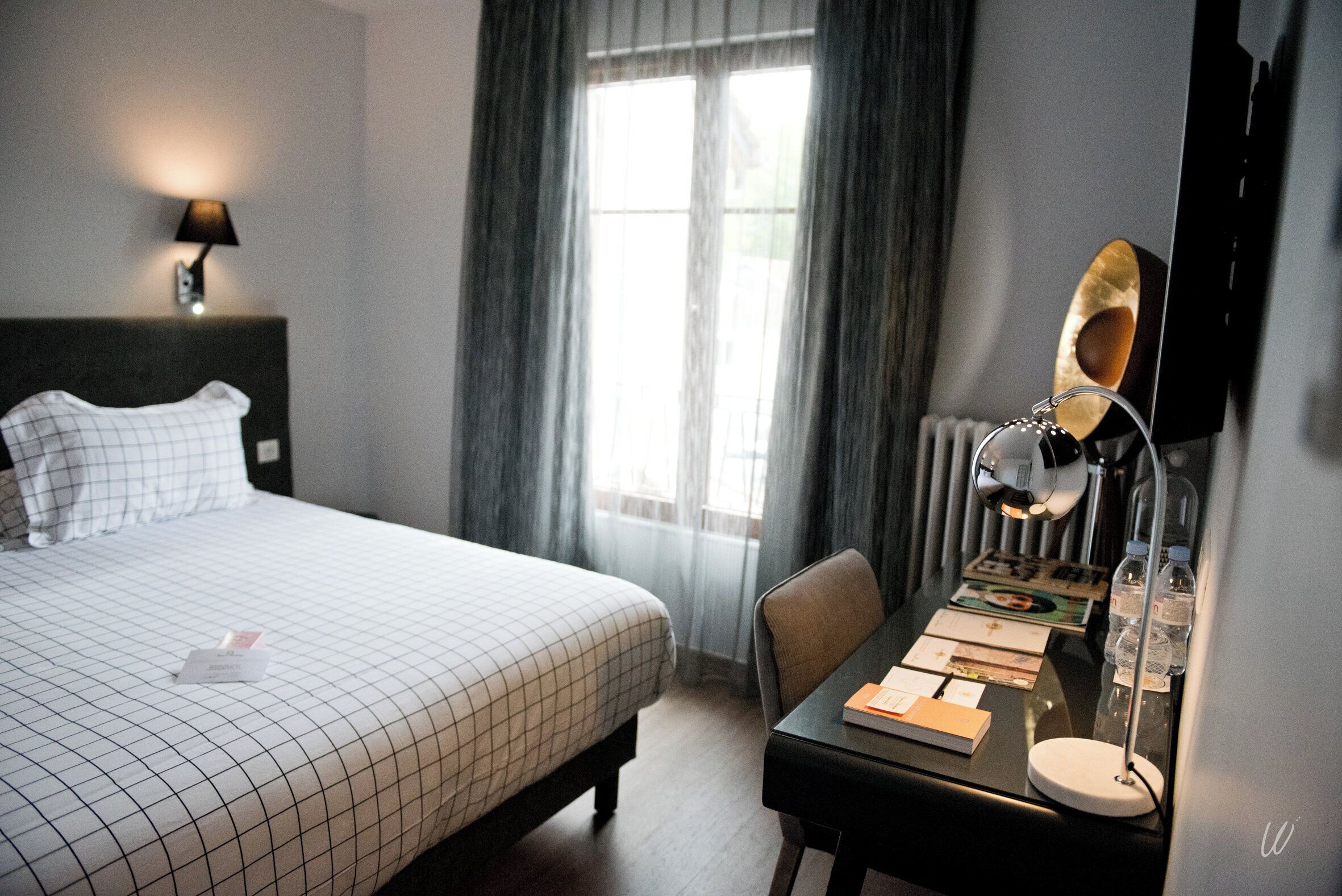 A hotel room with a bed, a desk, a lamp, and a window with curtains.