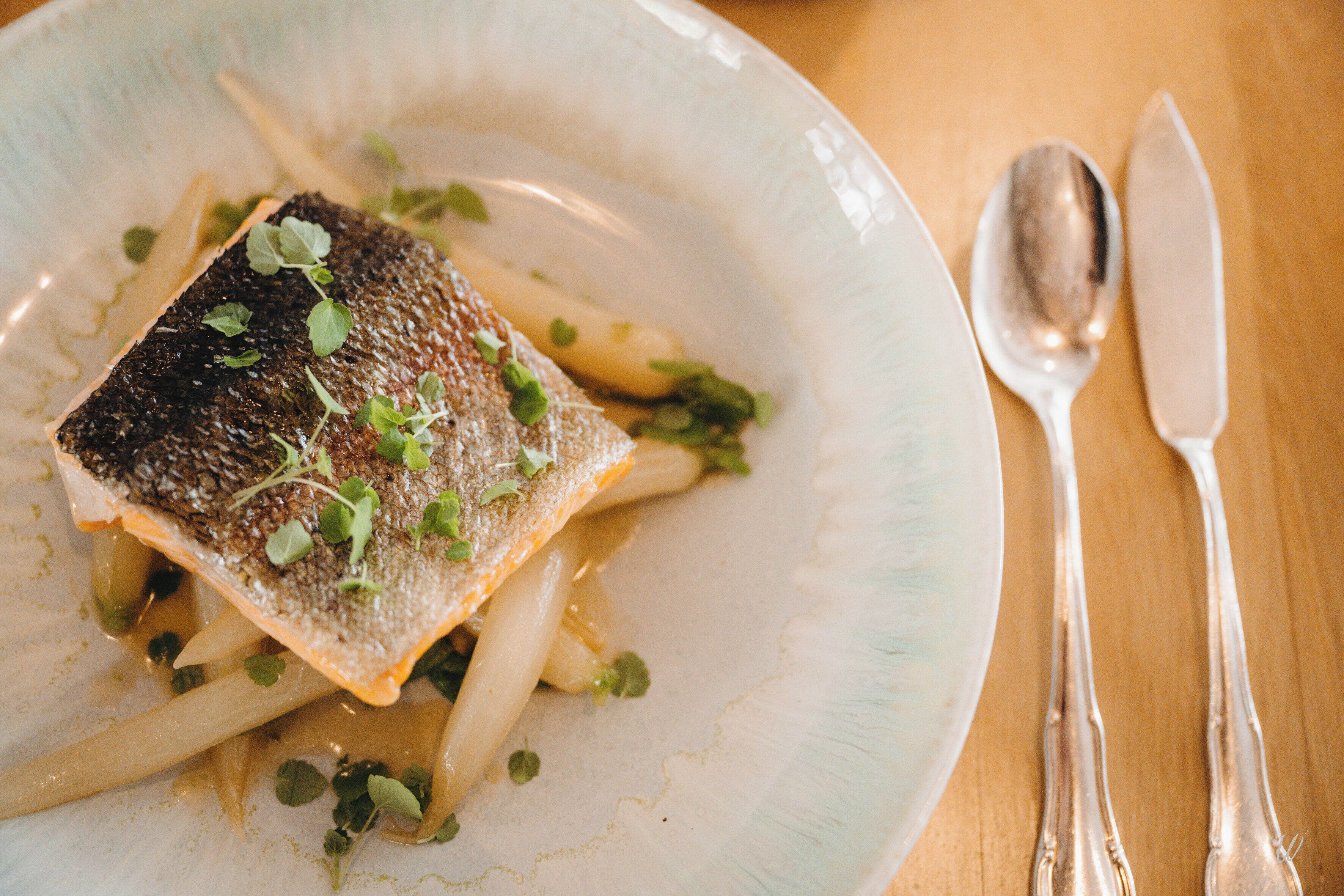 Une assiette contenant du poisson, des asperges et des légumes verts, posée sur une table avec une cuillère et un couteau.