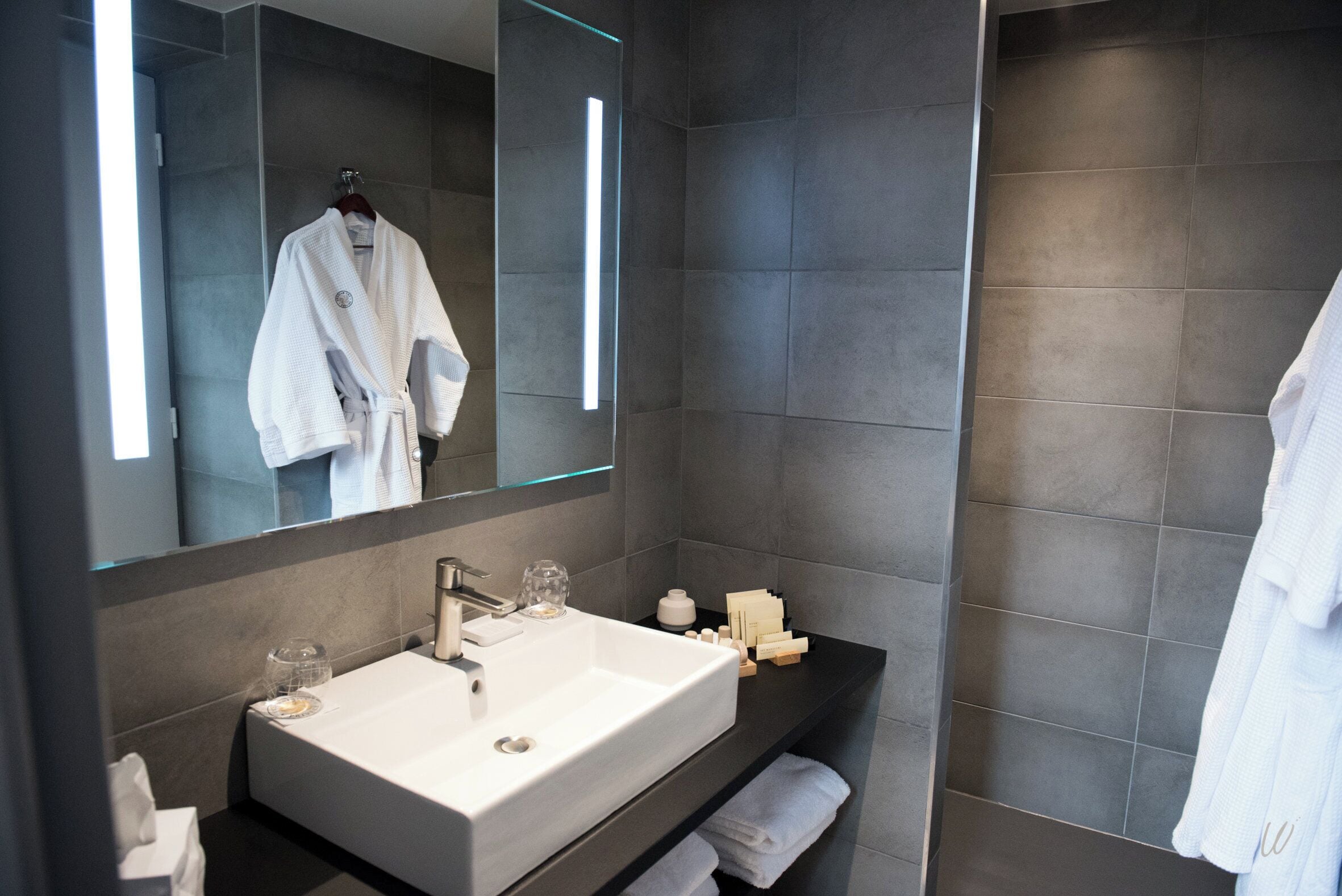 A modern bathroom with a large mirror, two sinks, and a dark tiled wall.