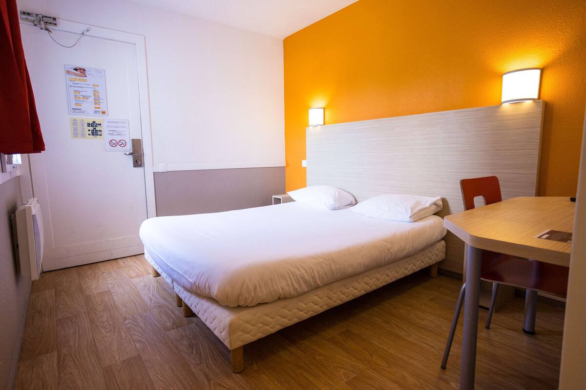 A hotel room with a bed, a table, and a chair. There is a door with a sign on it.