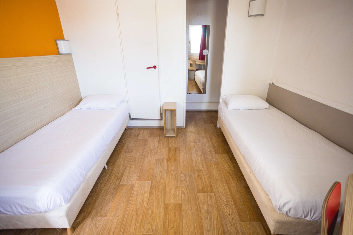A room with two single beds, a wooden floor, a white door, and a small wooden stool.