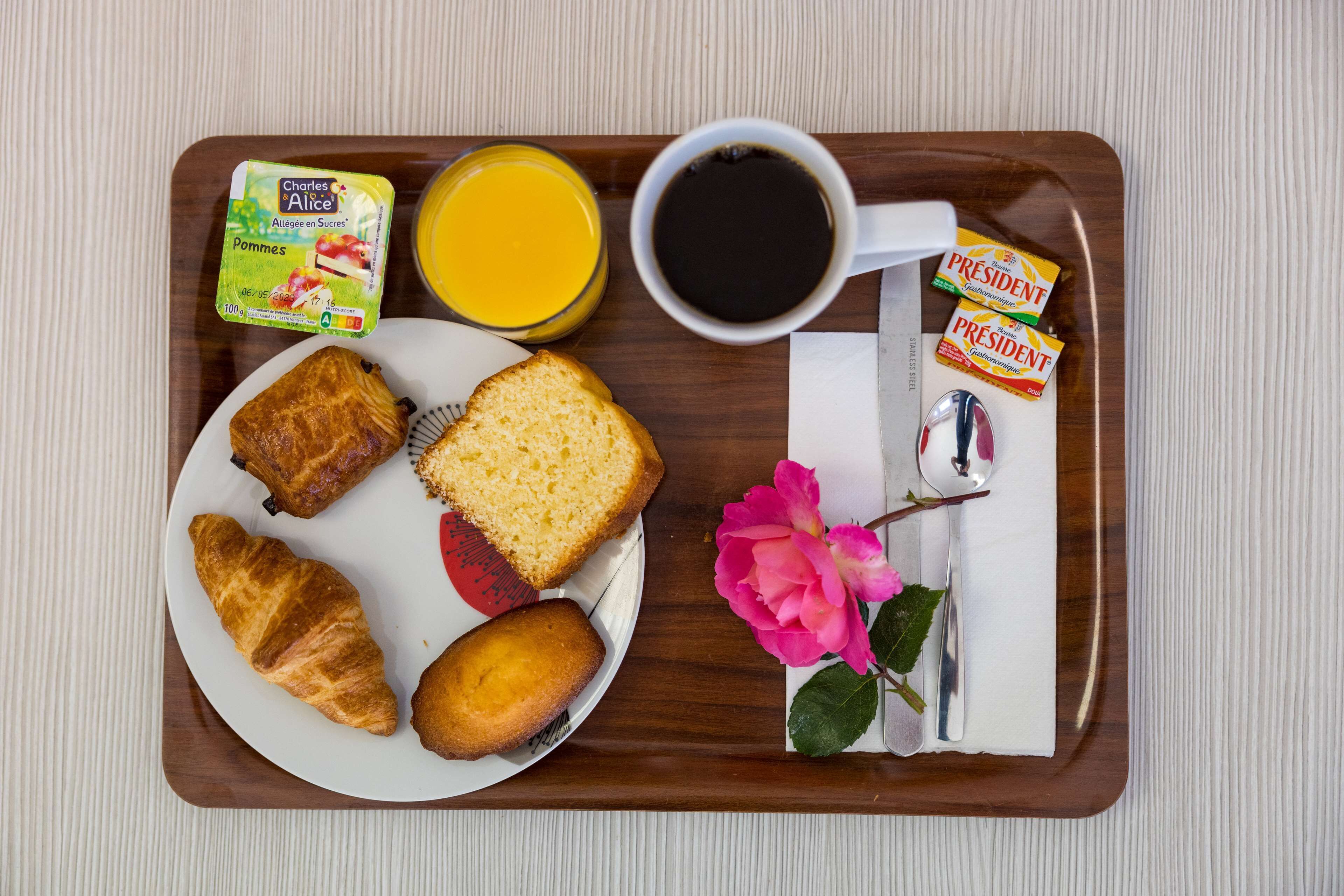 A breakfast tray with croissants, a slice of bread, a pastry, a glass of orange juice, a cup of coffee, a packet of butter, a packet of jam, a packet of sugar, a packet of tea, a rose, and a spoon.