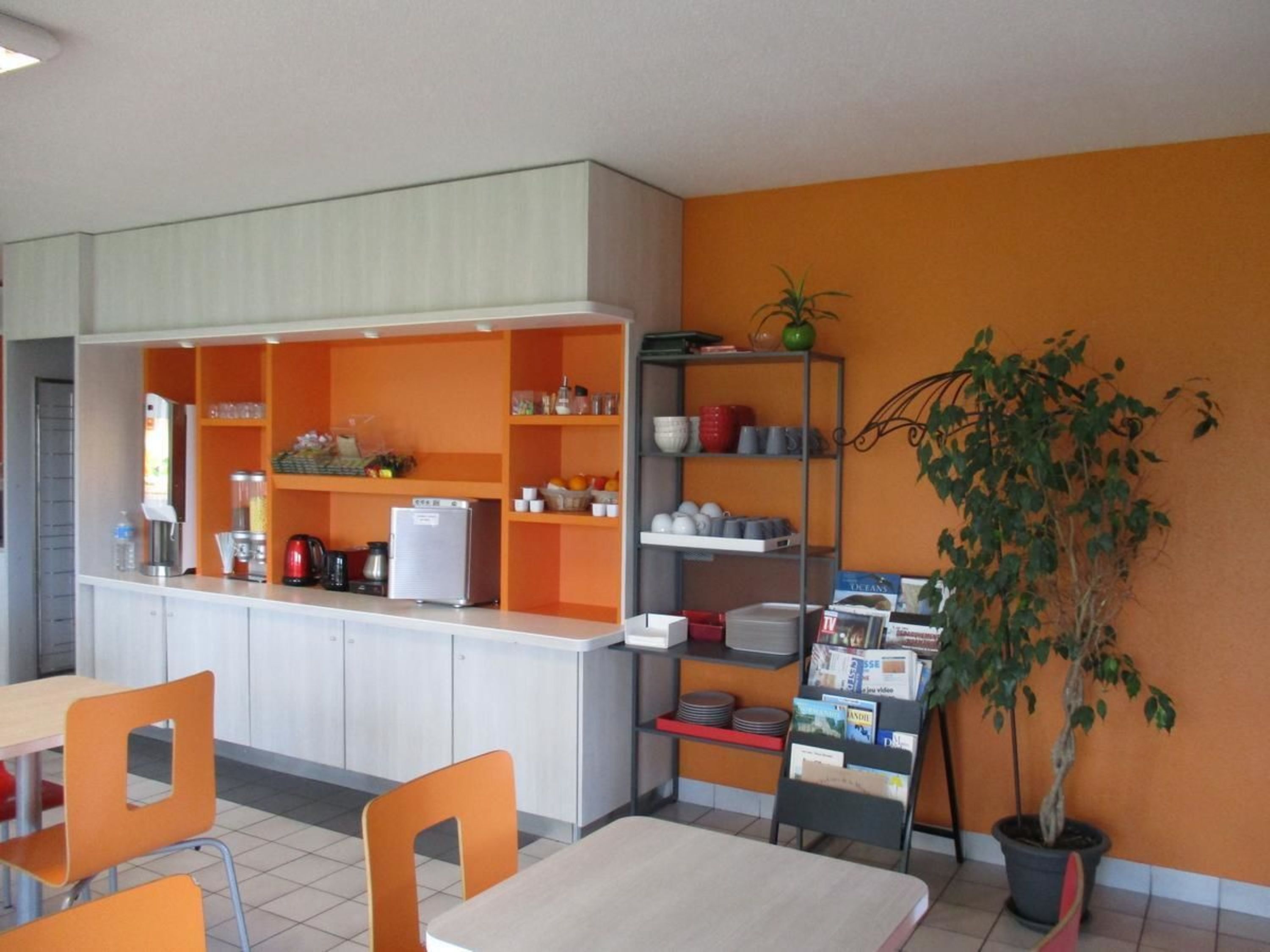 Eine moderne Küche mit orangenen Regalen, einer Kaffeemaschine und einem Kühlschrank. Orangene Stühle und ein Tisch sind vorhanden, außerdem ein Topf mit Pflanze.