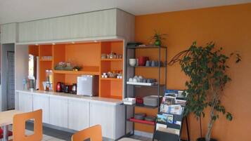 Una cocina moderna con estanterías naranja, una cafetera y un refrigerador. Hay sillas naranjas y una mesa, y se ve una planta en maceta.