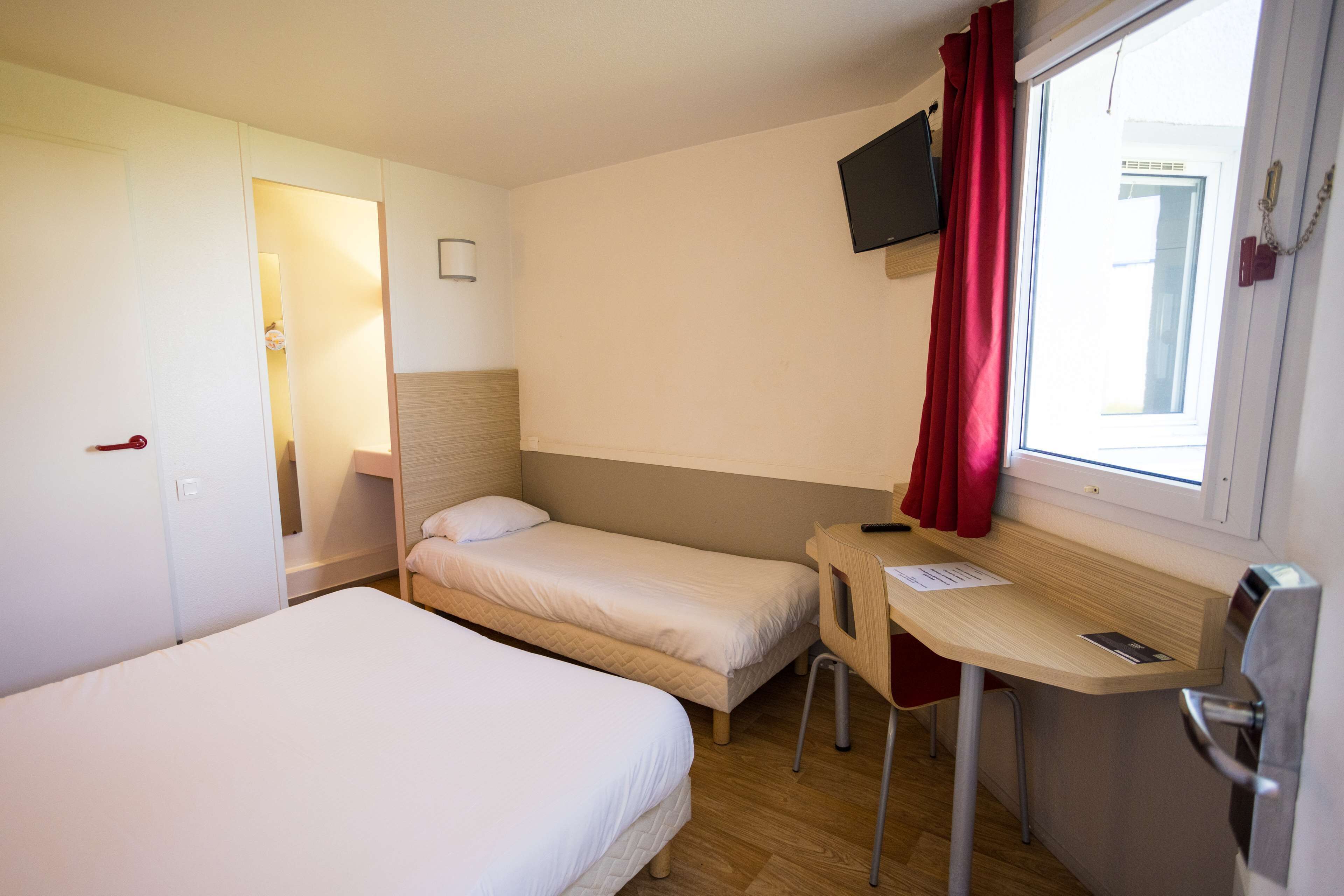 A hotel room with two single beds, a desk with a chair, a TV mounted on the wall, and a window with red curtains.