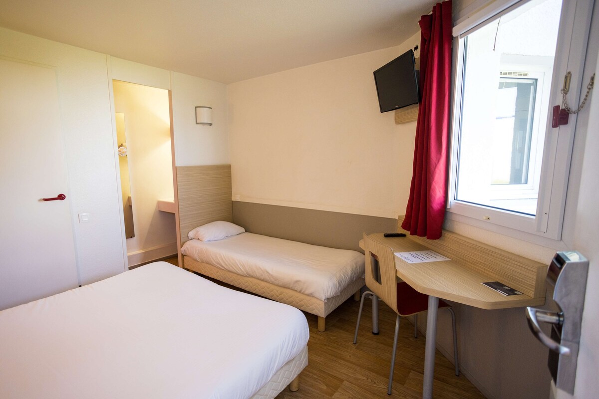 A hotel room with two single beds, a desk with a chair, a TV mounted on the wall, and a window with red curtains.
