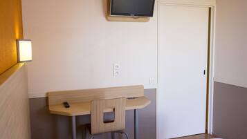 Una habitación de hotel con una cama individual, un escritorio pequeño con una silla, un televisor montado en la pared y una puerta.