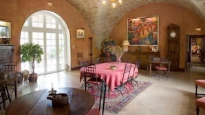 Un comedor amplio con una mesa redonda cubierta con un mantel rosa, rodeada de sillas. Se ve un ventanal grande que proporciona luz natural, y un cuadro en la pared.