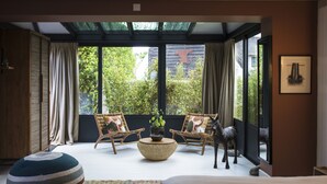 Une chambre moderne avec une grande fenêtre, deux fauteuils en osier, un pouf rond et un lit.
