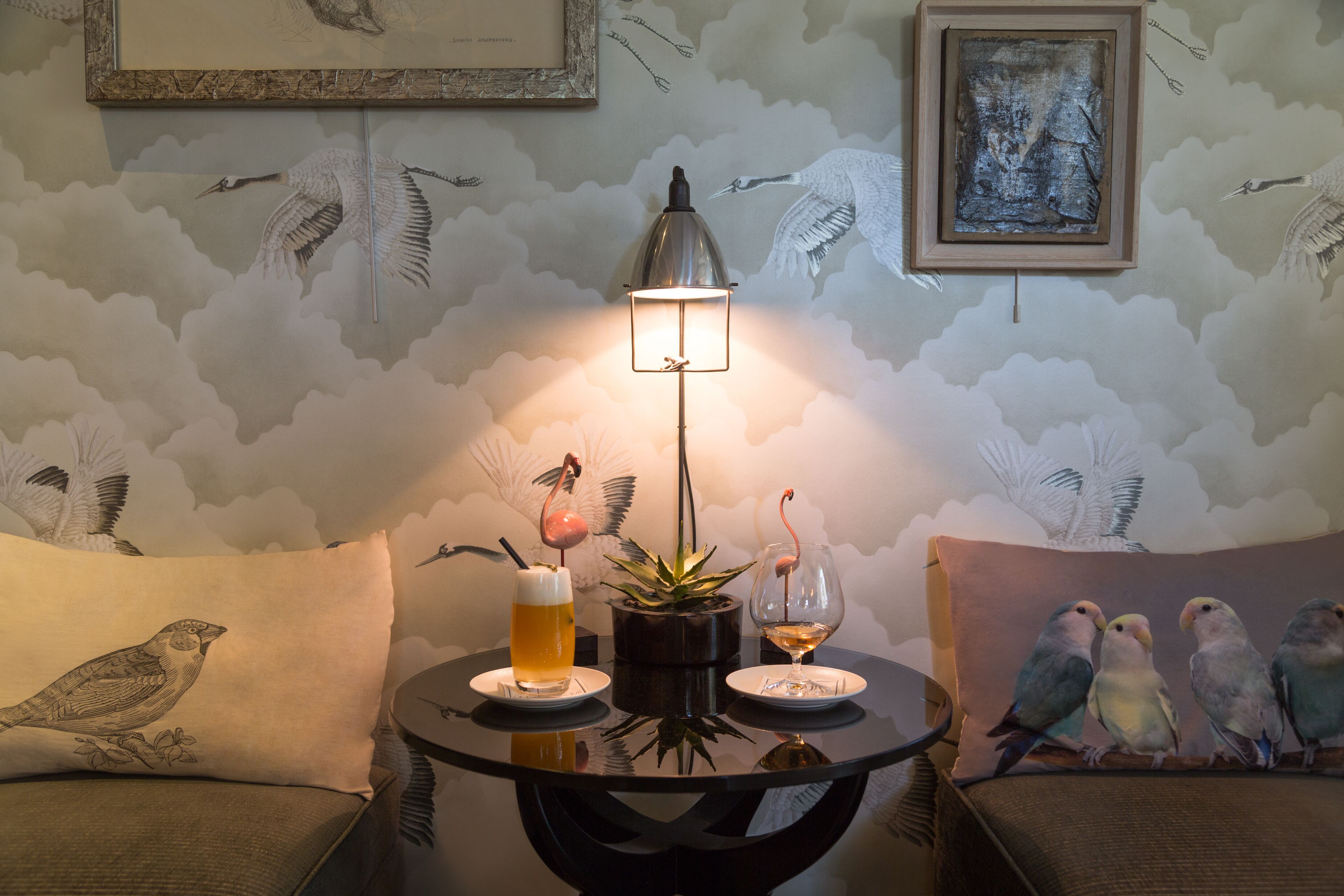 A round table with a lamp, two drinks, and a plant. The wall has bird murals.