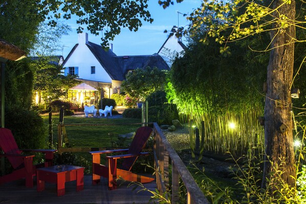 Eine Holzterrasse mit roten Stühlen und einem Tisch, im Hintergrund ein weißes Haus mit einem Reetdach und ein Bambuszaun.