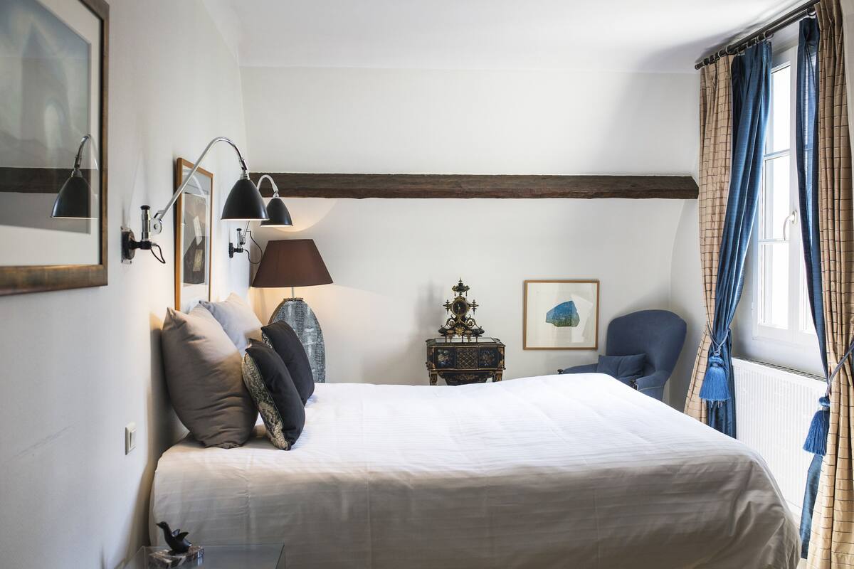 Une chambre à coucher avec un grand lit, une chaise, une petite table avec une lampe et une fenêtre avec des rideaux.