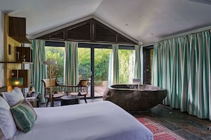 Een slaapkamer met een groot bed, een zithoek met stoelen, een ruime badkuip en uitzicht op groen door de ramen.