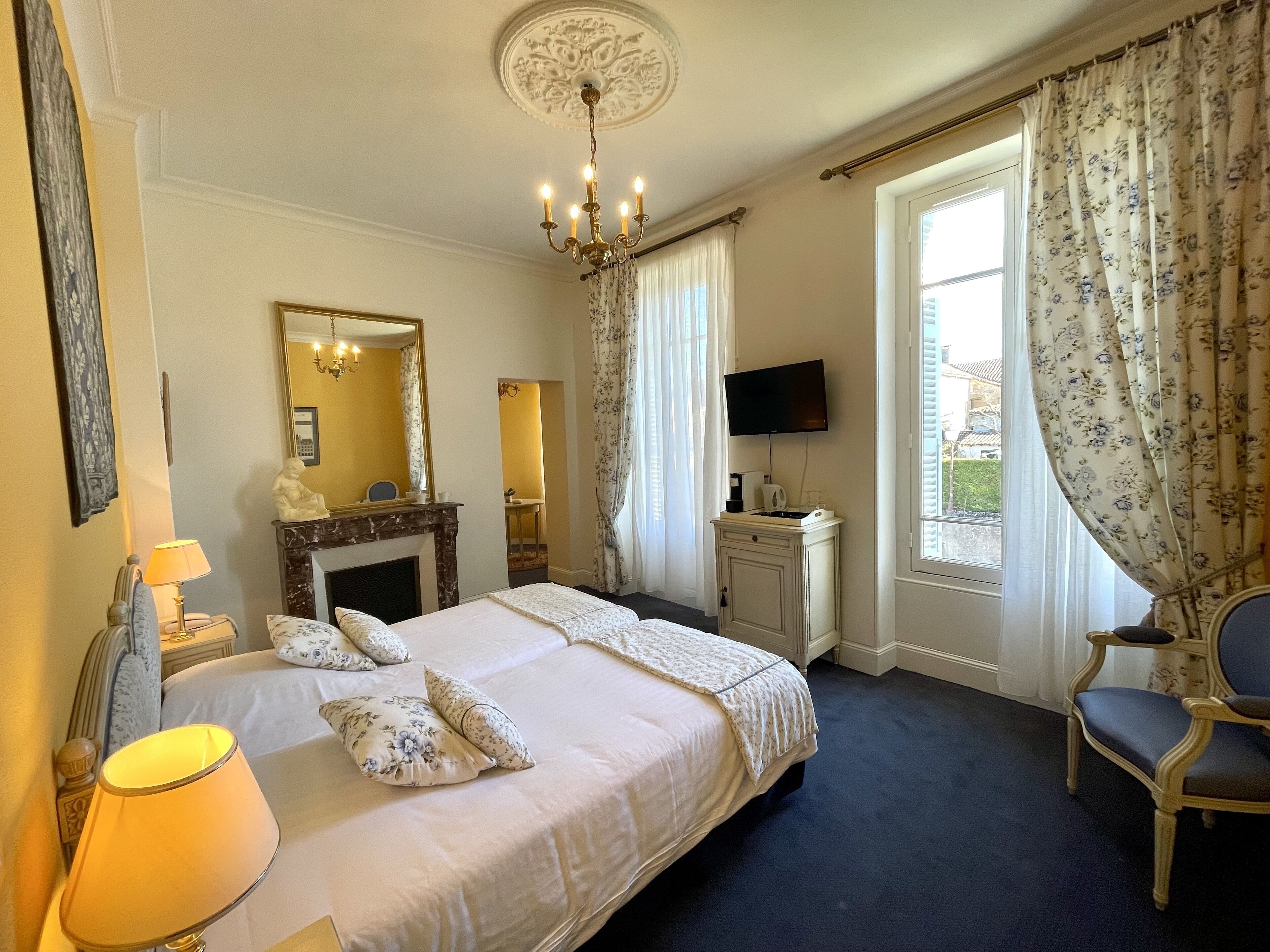 A hotel room with a large bed, a chandelier, a fireplace, a television, and a view of the outside.