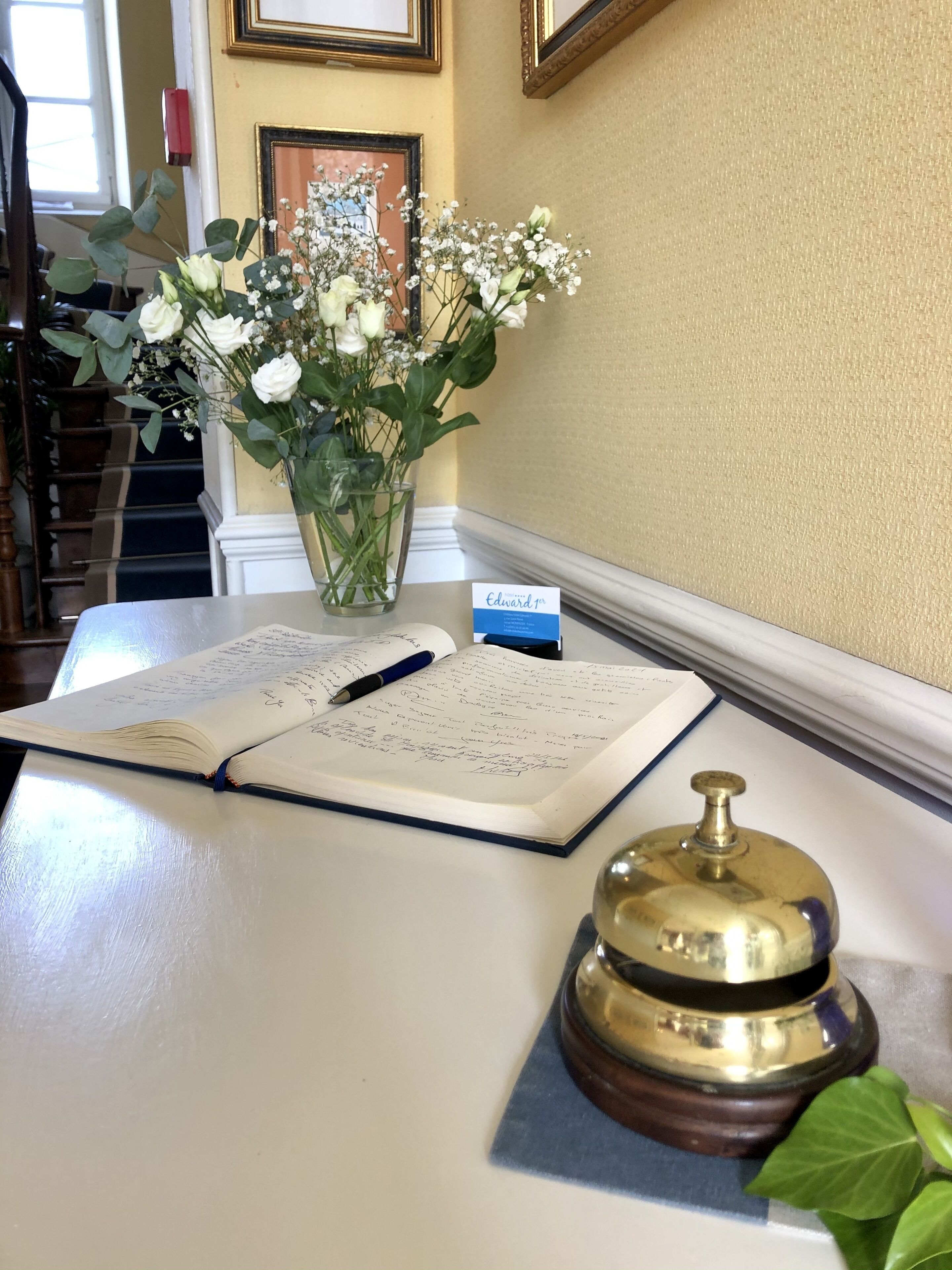 A desk with a notebook, pen, a brass bell, and a vase with flowers.