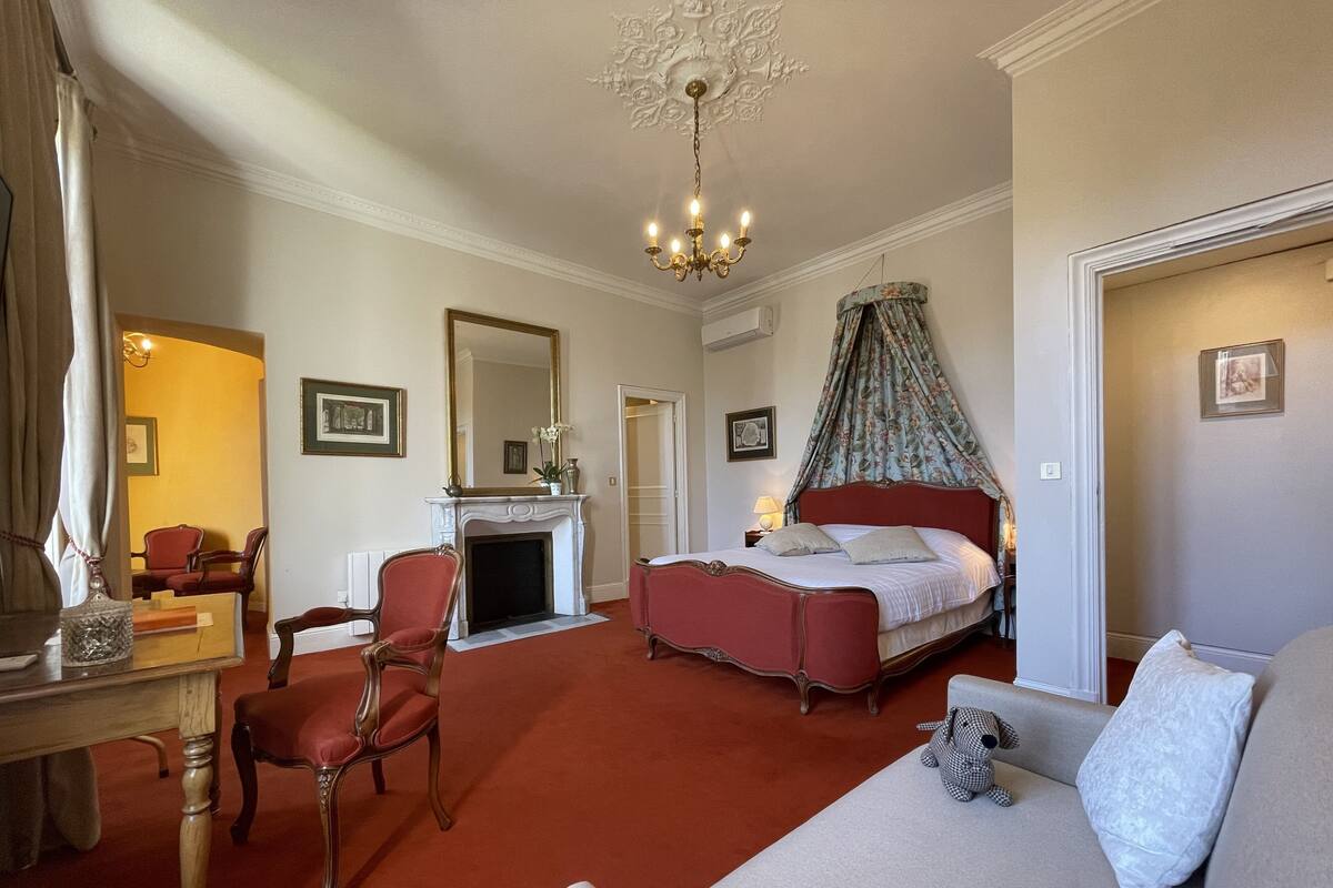 A hotel room with a bed, a sofa, a chair, a fireplace, a chandelier, and a mirror.