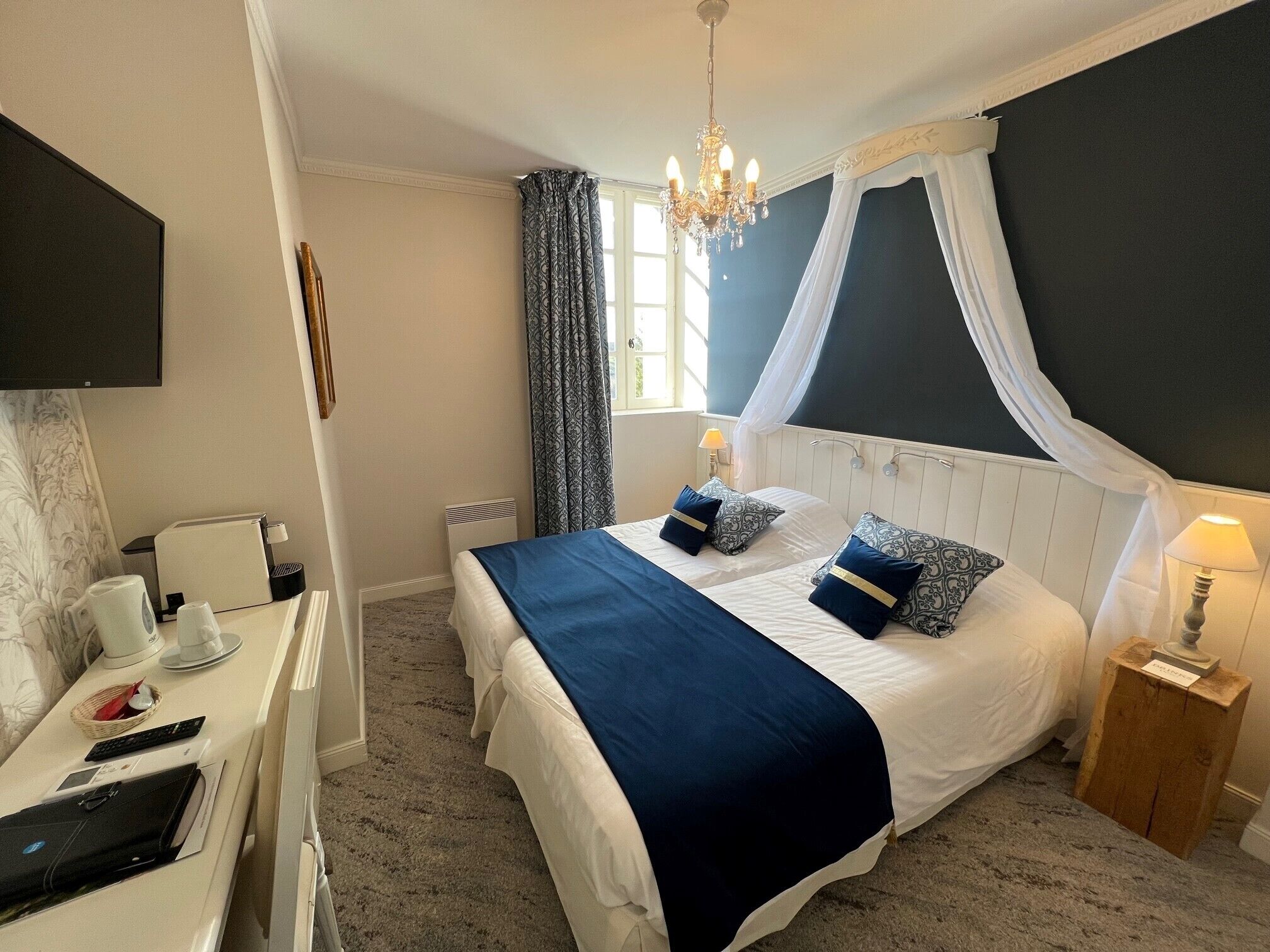 A bedroom with a bed, bedside tables, a desk with a chair, a television, and a chandelier.